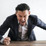 Managing Anxiety - Expressive angry businessman in formal suit looking at camera and screaming with madness while hitting desk with fist