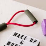 Healthy Weight - From above composition of dumbbells and massage double ball and tape and tubular expanders surrounding light box with wake up and workout words placed on white surface of table