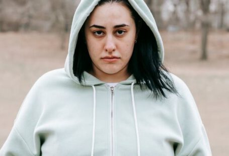 Weight Maintenance - Sad plus sized female in hoodie touching and showing fat on belly while standing in leafless park and looking at camera