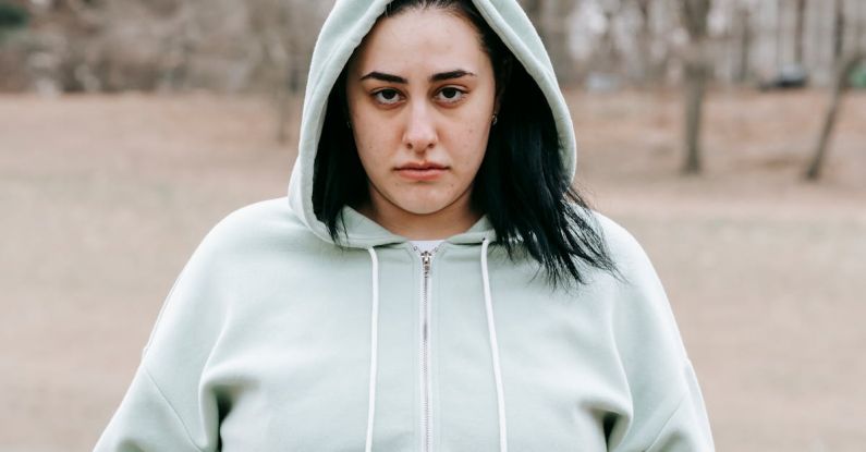 Weight Maintenance - Sad plus sized female in hoodie touching and showing fat on belly while standing in leafless park and looking at camera